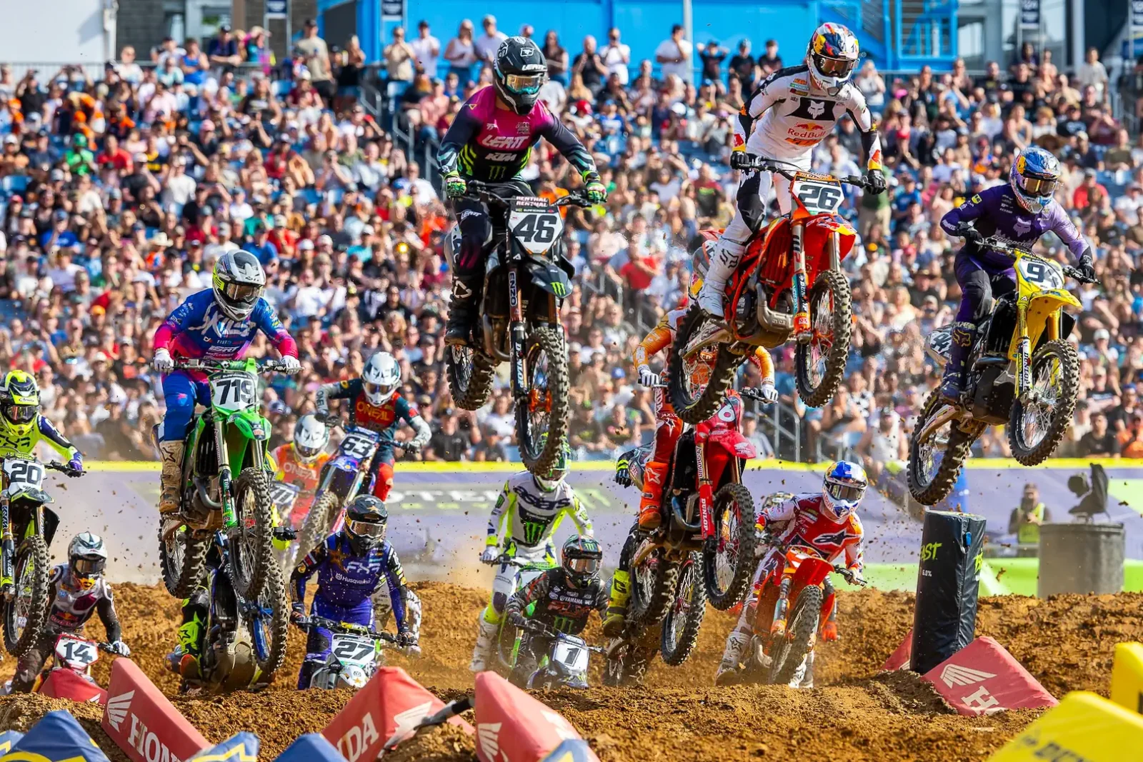 Detalhe de um piloto de SuperMotocross em ação, saltando sobre um obstáculo com a moto no ar, em um estádio lotado.