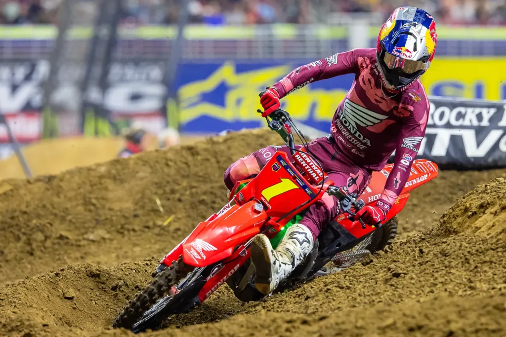 Detalhe de um piloto de SuperMotocross em ação, saltando sobre um obstáculo com a moto no ar, em um estádio lotado.