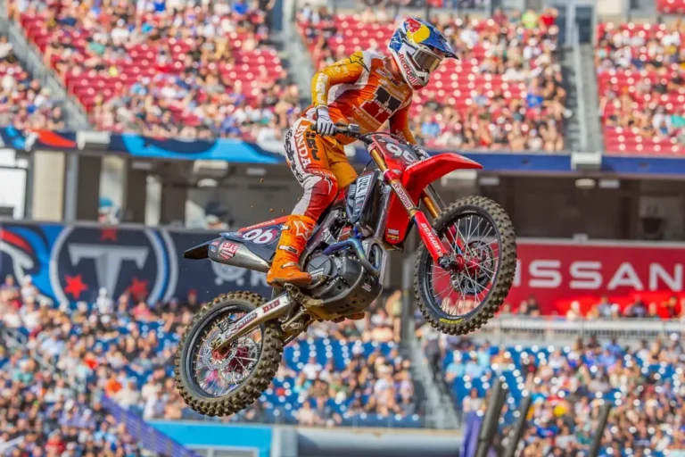 Detalhe de um piloto de SuperMotocross em ação, saltando sobre um obstáculo com a moto no ar, em um estádio lotado.