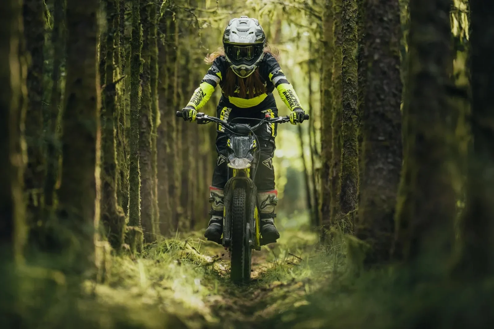 Triumph TXP 20 em ação, em um cenário de trilha com obstáculos naturais, exibindo sua agilidade e design específico para trials.