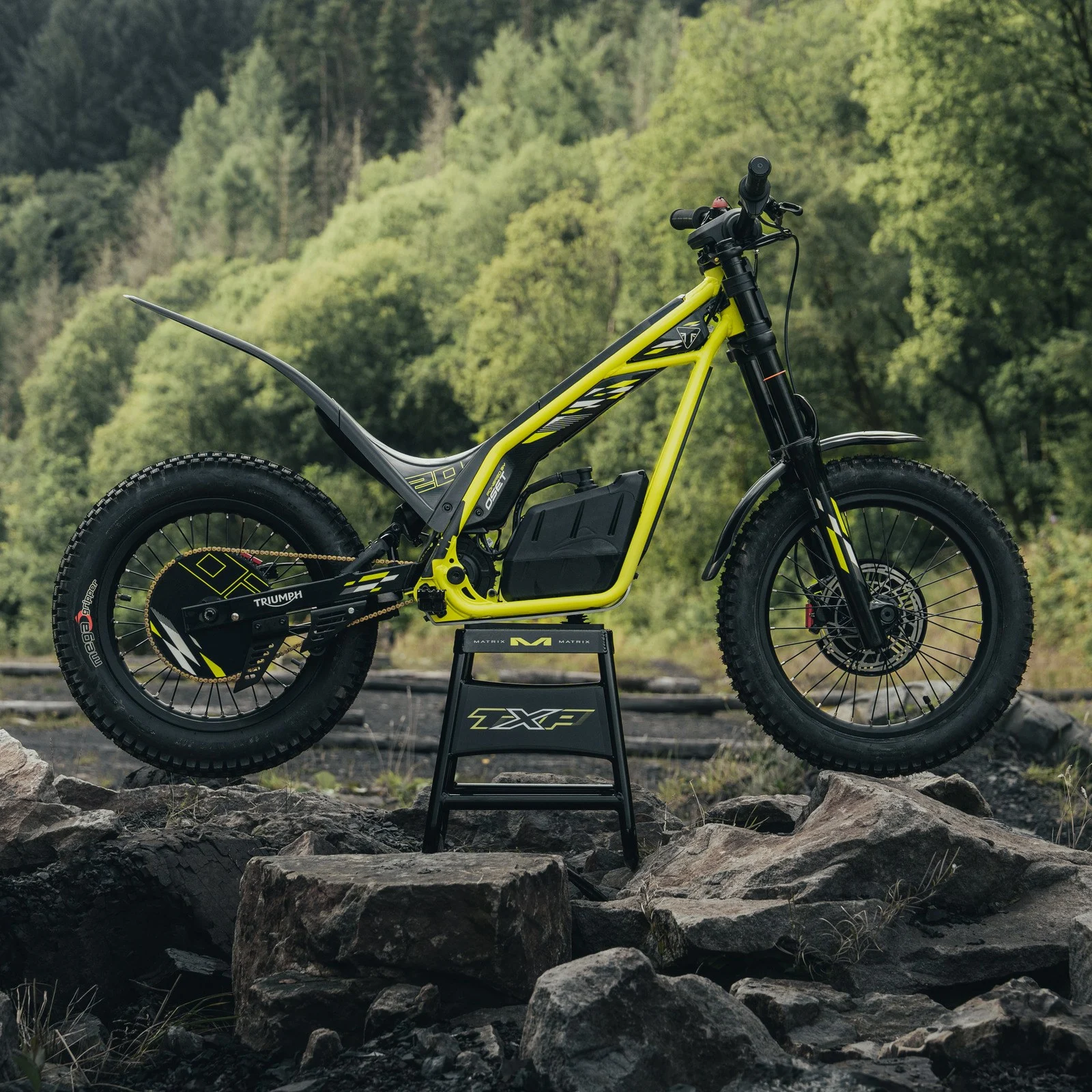 Triumph TXP 20 em ação, em um cenário de trilha com obstáculos naturais, exibindo sua agilidade e design específico para trials.