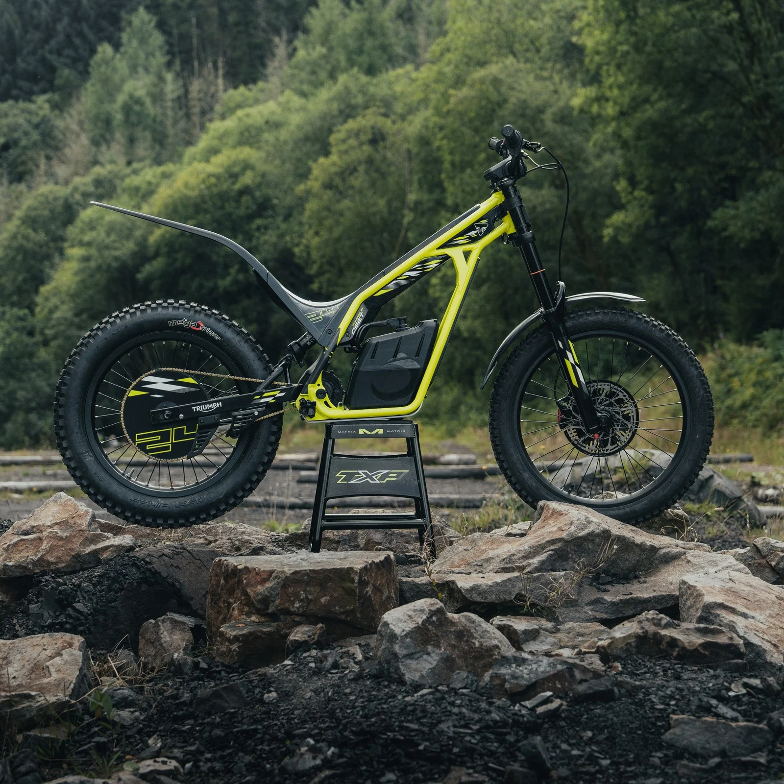 Triumph TXP 20 em ação, em um cenário de trilha com obstáculos naturais, exibindo sua agilidade e design específico para trials.