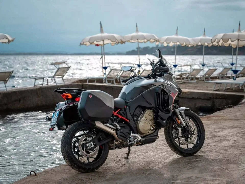 Ducati Multistrada V4 S Grand Tour 2024 em vermelho com malas laterais e detalhes em preto, estacionada em estrada europeia.